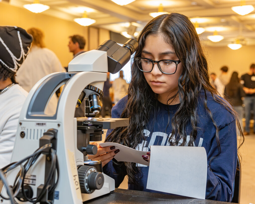 female student with eye glasses holding a paper in front of a microscope